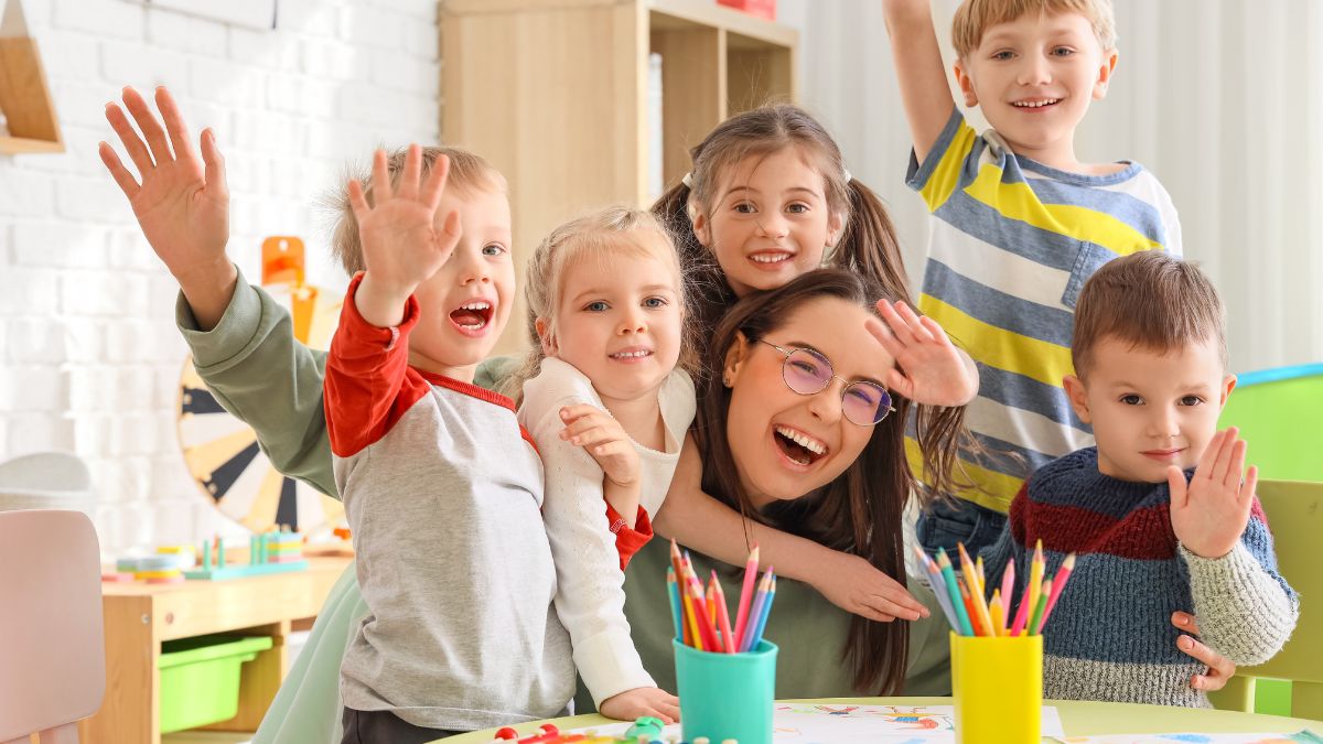 Kindergartenkinder und Pädagogin winken fröhlich in einem hellen Gruppenraum